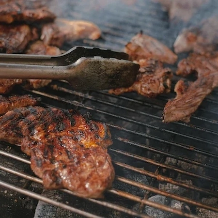 steaks being cooked on a grill with silver tongs on the side.