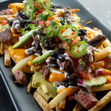 nacho fries on a black plate with toppings like steak, cheese, avocado, black olives, tomatoes and jalapenos