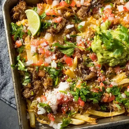 a silver pan of nacho fries with nacho toppings like tomatoes, ground beef, onions, guacamole, and cilantro.