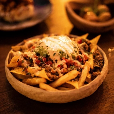 a wooden bowl with nacho fries in it topped with seasoning, ground beef, sour cream, salsa, guacamole, and cheese